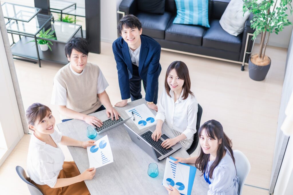 明るいオフィスで、ノートパソコンと資料を前に打ち合わせをしている5人のビジネスパーソン